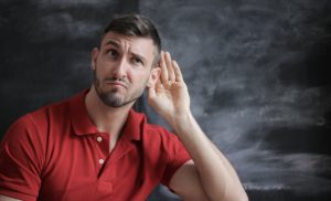 Man in red t-shirt frowning with hand up to ear like he's listening