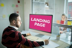 Man sitting at computer desk looking a screen with landing page written in big letters on a pink background.