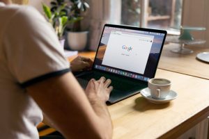 Showing man sitting at desk on computer with Google search page on the screen and a cup of coffee next to him. 